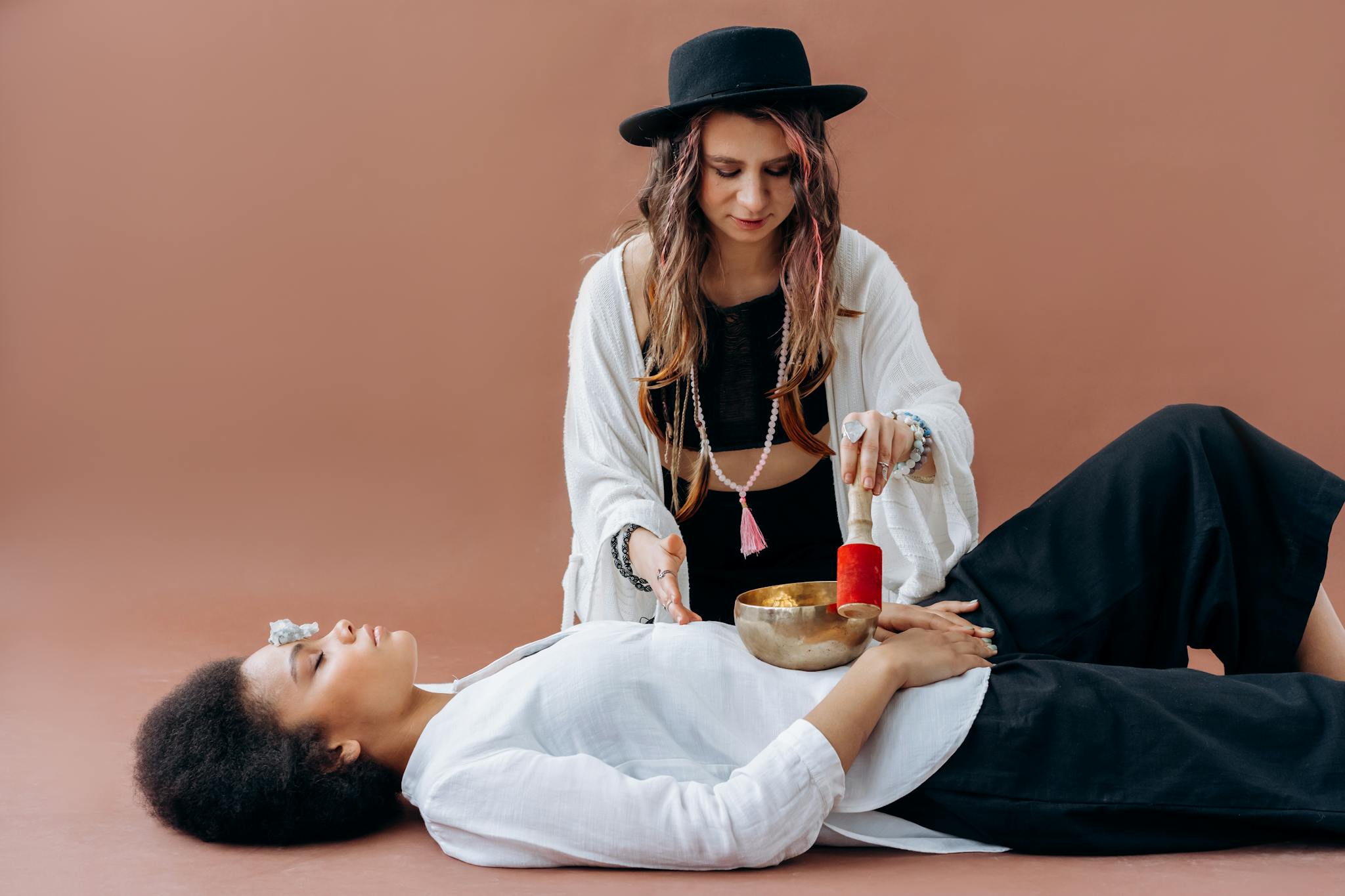 Woman practicing sound healing with singing bowl over another woman. Meditative and relaxing wellness session.